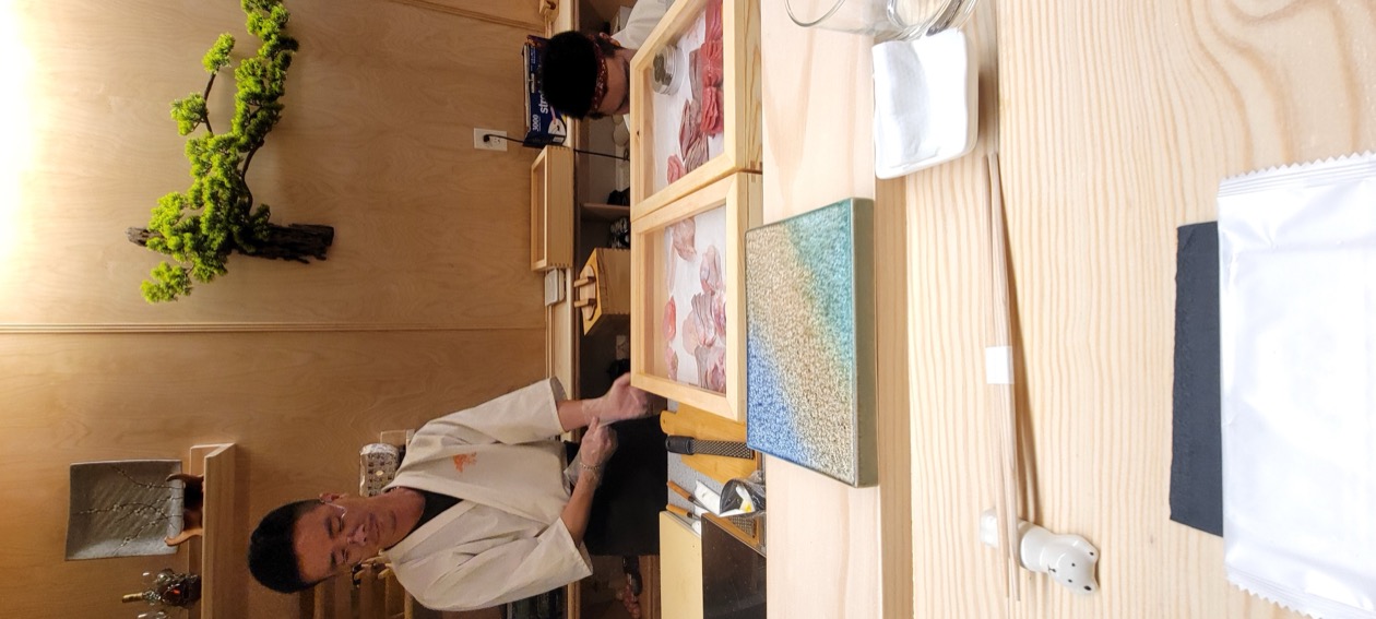 Omakase counter with the chef preparing fish, bonsai tree visible on the shelf behind